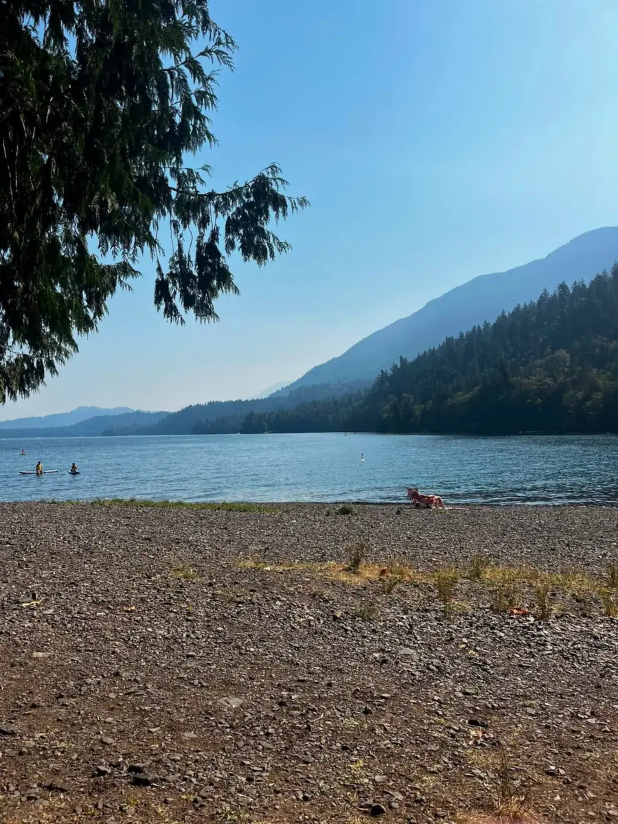 Cultus Lake seen from a stony shore, forested mountains folding into blue haze on the far side and two paddleboarders on flat water