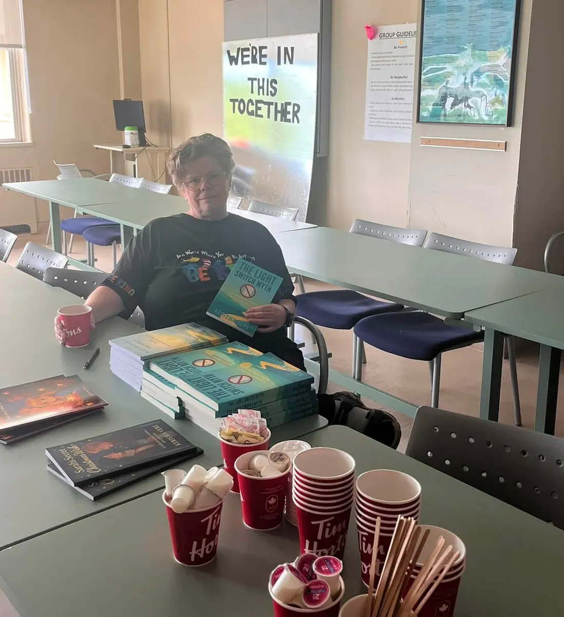 Kerisma Vere sitting at a long table in a community group room with stacks of her books, a coffee station set out, and a hand-lettered We're In This Together sign on the wall behind her