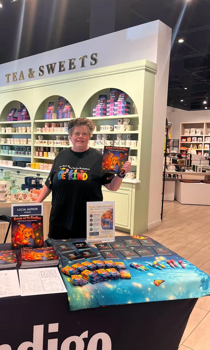 Kerisma Vere standing behind a book-signing table at an Indigo bookstore, holding up a copy of Krizzy and the Fireflies beside a Local Author Signing sign and stacks of her books